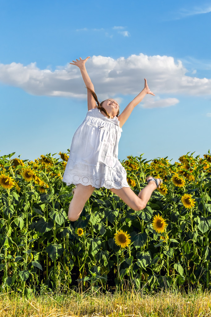 Girl in white sun-dress jumping on a background of a field of yellow sunflowers and blue sky